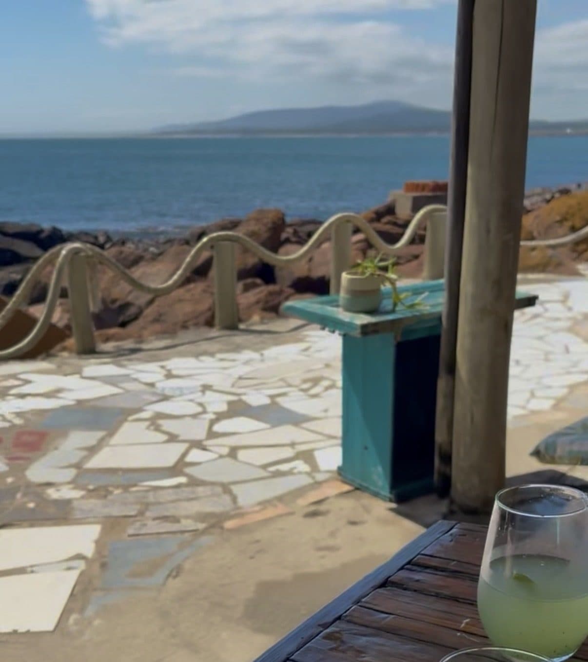 Terraço do Puertito Don Anselmo sobre as pedras de Punta Fría, com o mar e os morros ao fundo e uma limonada na mesa